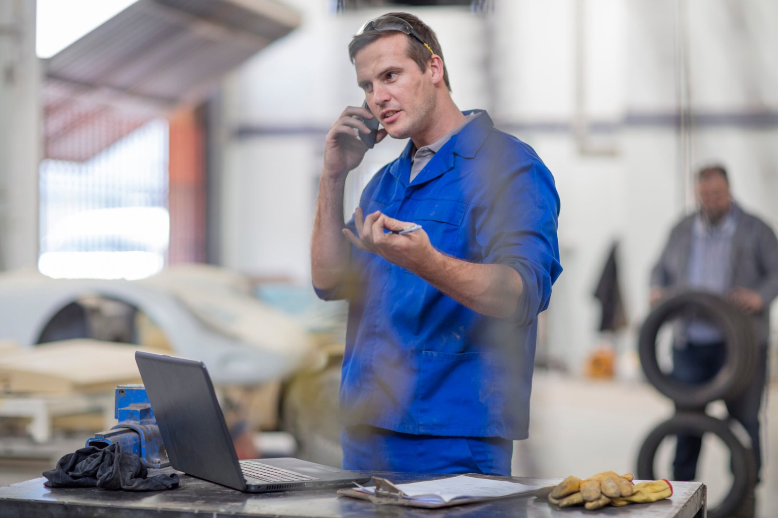 Mechanic talking on phone in a workshop with a laptop and car in background.